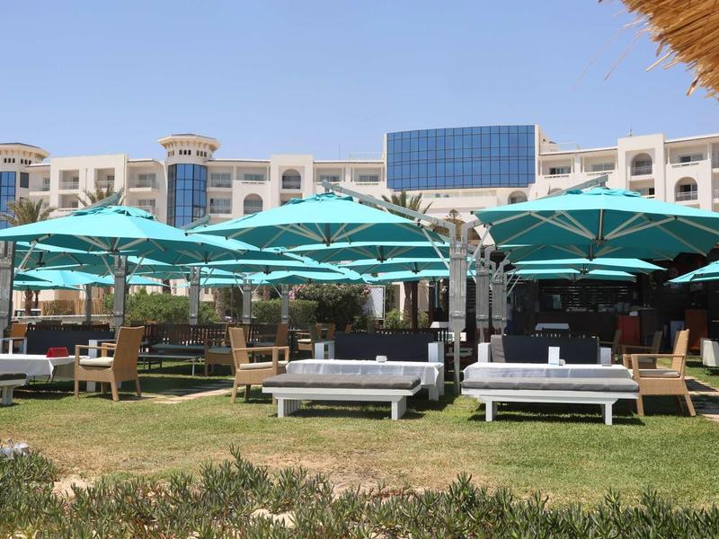 Ogród hotelowy z leżakami i turkusowymi parasolami przed dużym białym budynkiem.