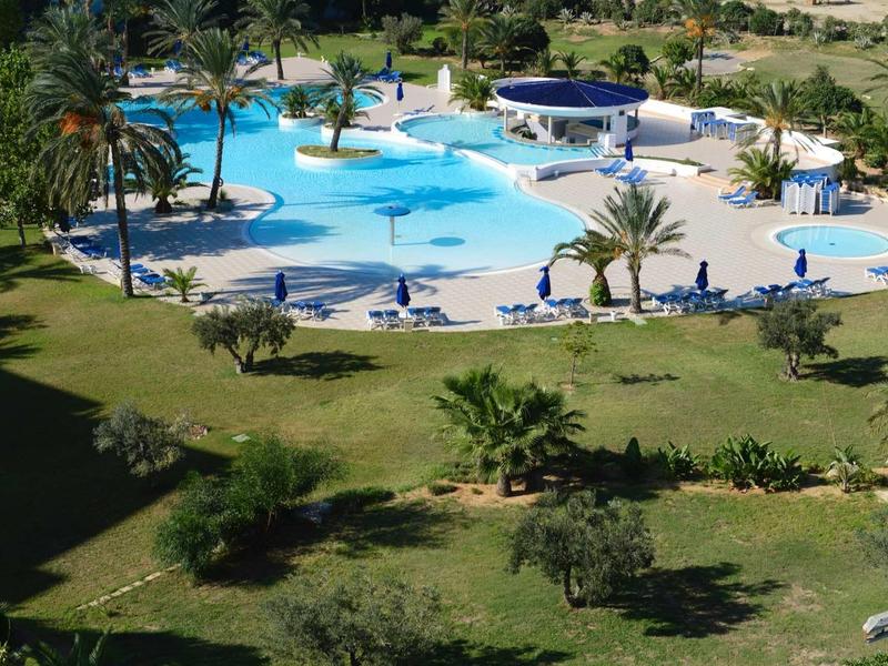 Duży basen hotelowy z niebieską wodą, leżakami i palmami na zielonym trawniku