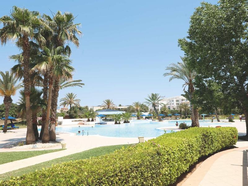 Duży basen z palmami i miejscami do siedzenia w słonecznym ośrodku hotelowym.