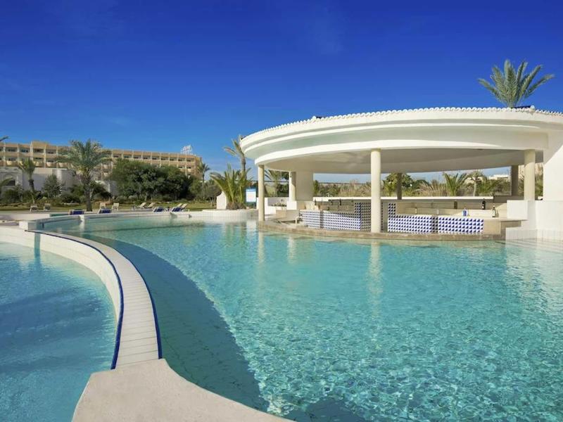 Duży basen z białym okrągłym pawilonem i palmami pod jasnym niebieskim niebem.