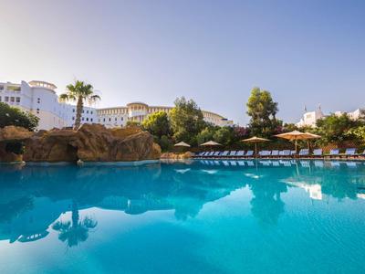 Großer blauer Pool mit Liegestühlen, Felsen und Palmen vor weißen Hotelgebäuden bei klarem Himmel.