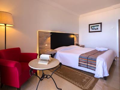 Nowoczesny pokój hotelowy z podwójnym łóżkiem, czerwonym fotelem, lampą stojącą i małym stolikiem z książkami.