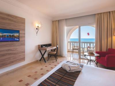 Jasny pokój hotelowy z łóżkiem, czerwonym fotelem, biurkiem i balkonem z widokiem na morze.