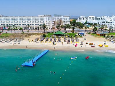 Widok z lotu ptaka na duży hotel przy plaży z białymi budynkami, piaszczystą plażą, pomostem i turkusowym morzem.