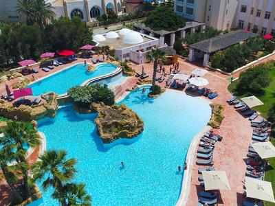 Duży basen hotelowy z parasolami, leżakami i otoczony zielenią.