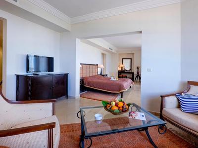 Elegancki pokój hotelowy z częścią wypoczynkową, szklanym stołem, miską owoców i widokiem na wygodne łóżko.