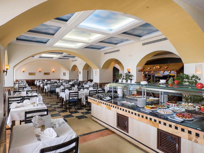 Przestronna sala jadalna hotelu z bufetem i schludnie nakrytymi stołami z białymi obrusami.