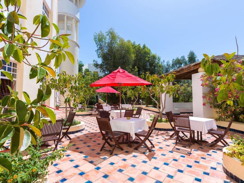 Słoneczne patio z drewnianymi krzesłami, stołem i czerwonym parasolem otoczone roślinami.