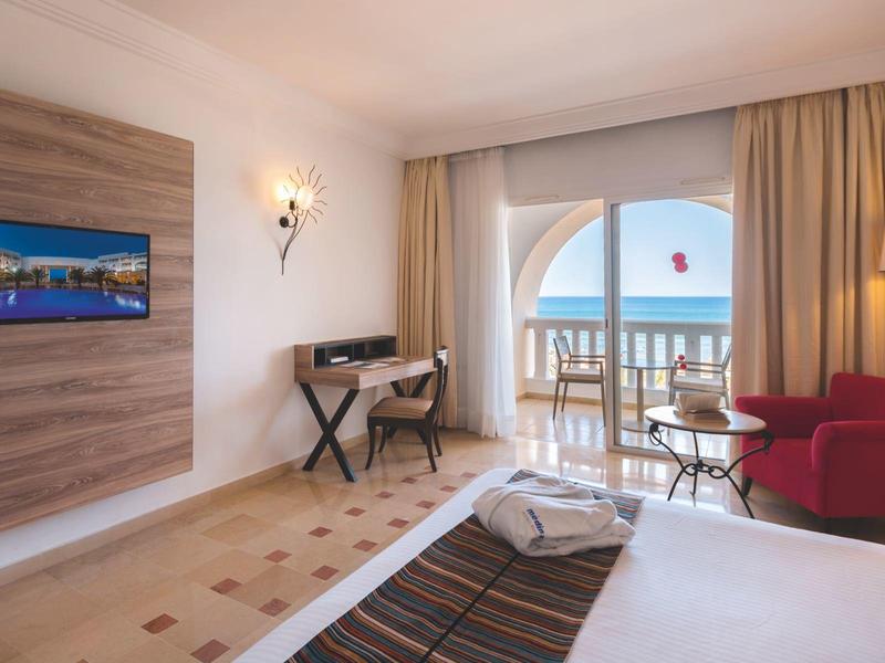 Jasny pokój hotelowy z łóżkiem, czerwonym fotelem, biurkiem i balkonem z widokiem na morze.