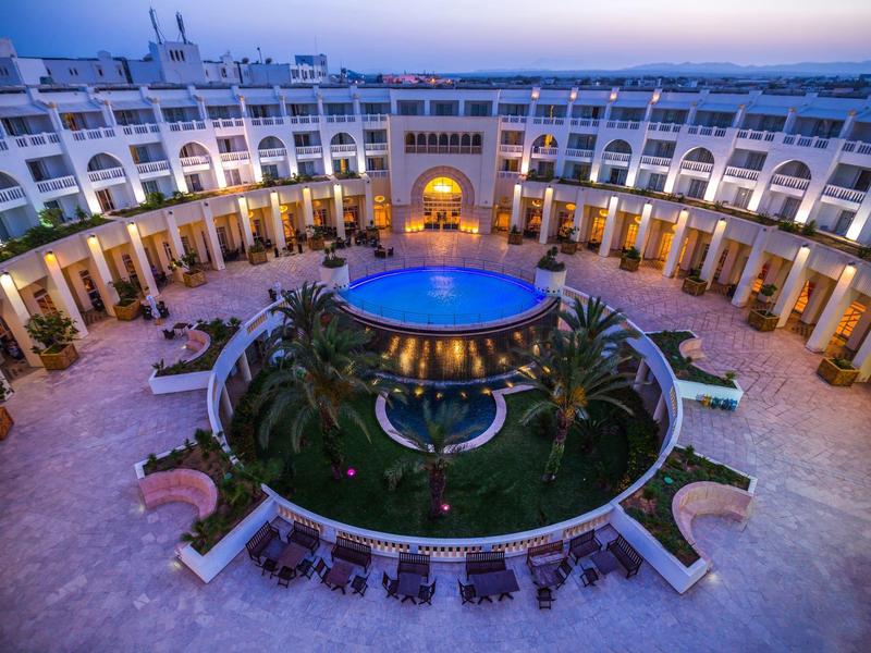 Okrągły hotel z oświetlonym basenem i palmami w centrum, otoczony balkonami pokoi.