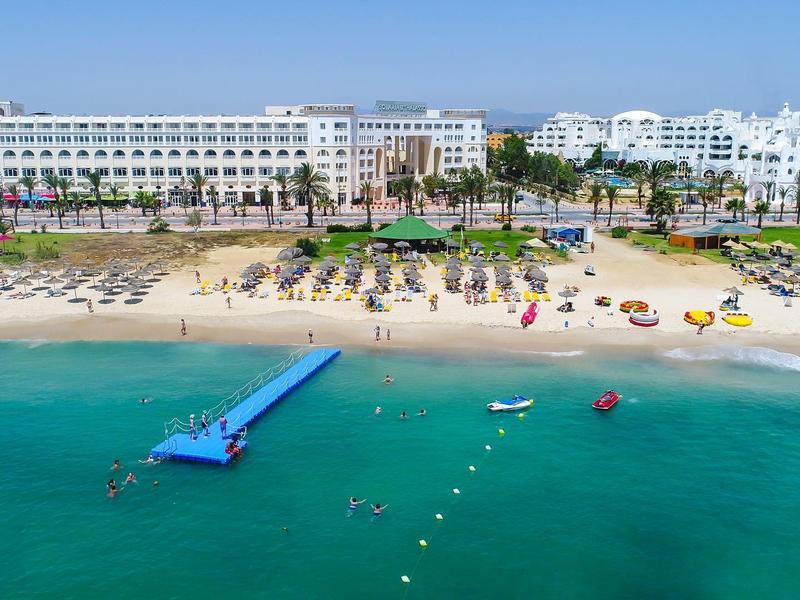 Widok z lotu ptaka na duży hotel przy plaży z białymi budynkami, piaszczystą plażą, pomostem i turkusowym morzem.