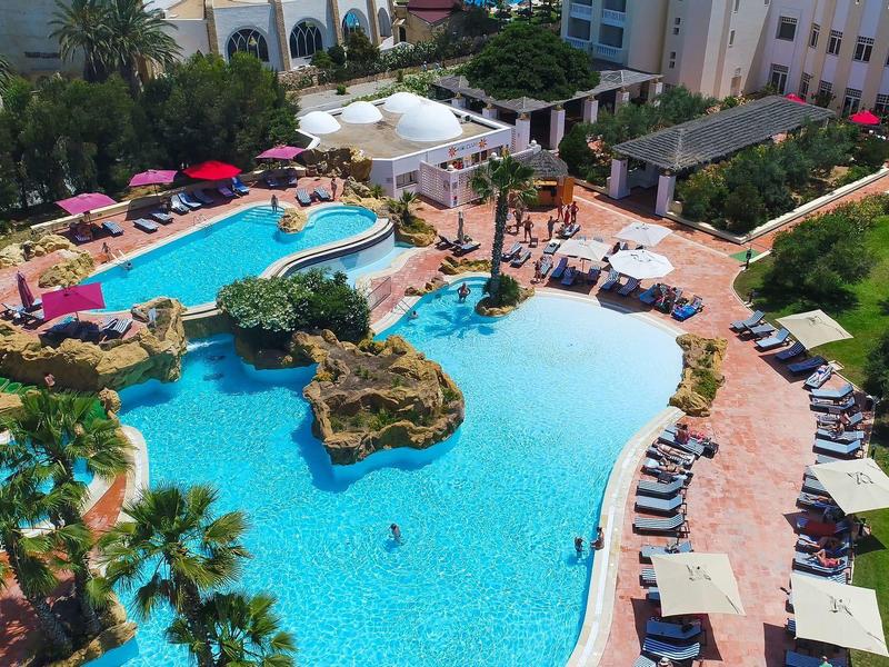 Duży hotelowy basen z wyspą, otoczony leżakami, parasolami i zielenią.