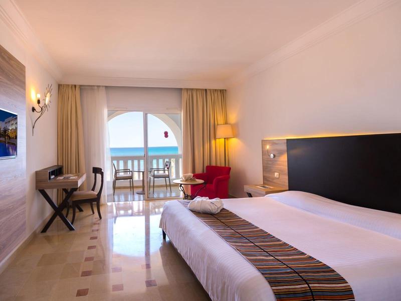 Jasny pokój hotelowy z dużym łóżkiem, czerwonym fotelem, biurkiem i balkonem z widokiem na morze.