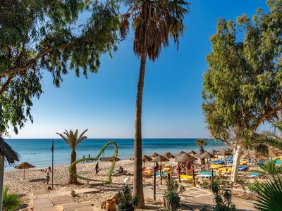Strand met parasols, ligstoelen en bomen onder een helderblauwe lucht en zee.