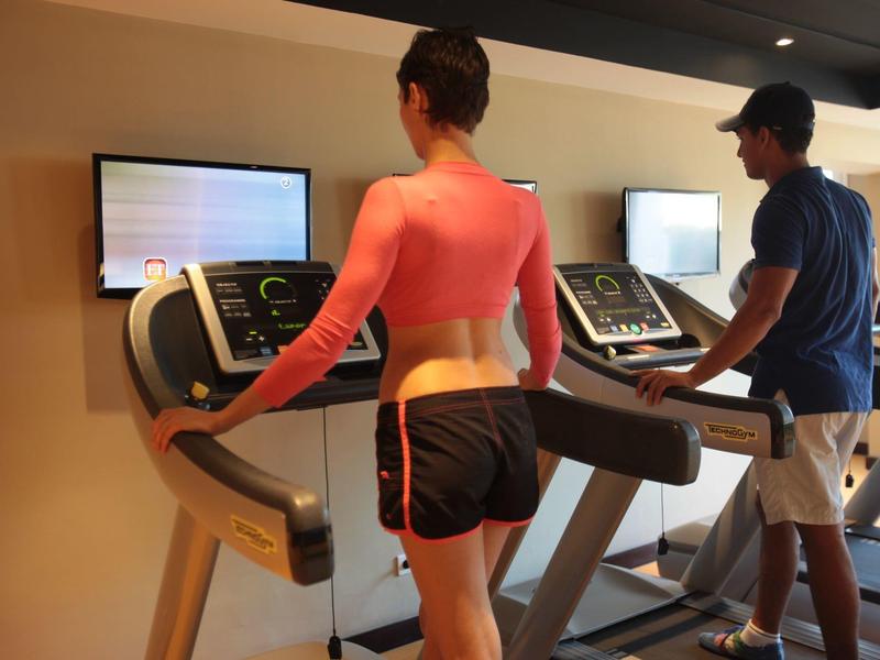 Dwie osoby ćwiczą na bieżniach w siłowni z monitorami na ścianie.