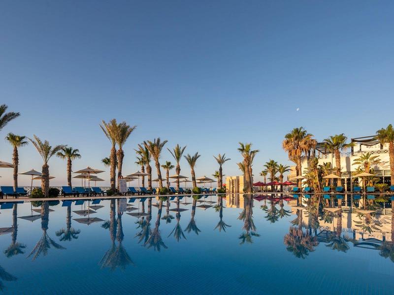 Basen hotelowy z palmami i leżakami pod jasnym, niebieskim niebem.