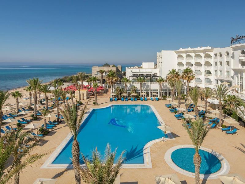 Duży basen hotelowy z leżakami i widokiem na morze pod błękitnym niebem.