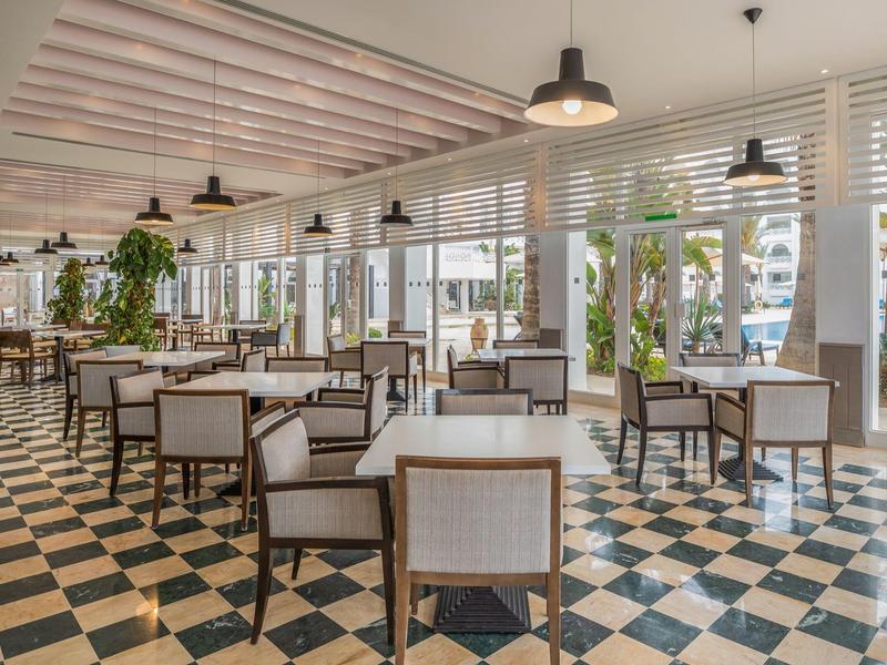 Nowoczesna, jasna restauracja z klasycznymi czarno-białymi płytkami i wieloma roślinami.