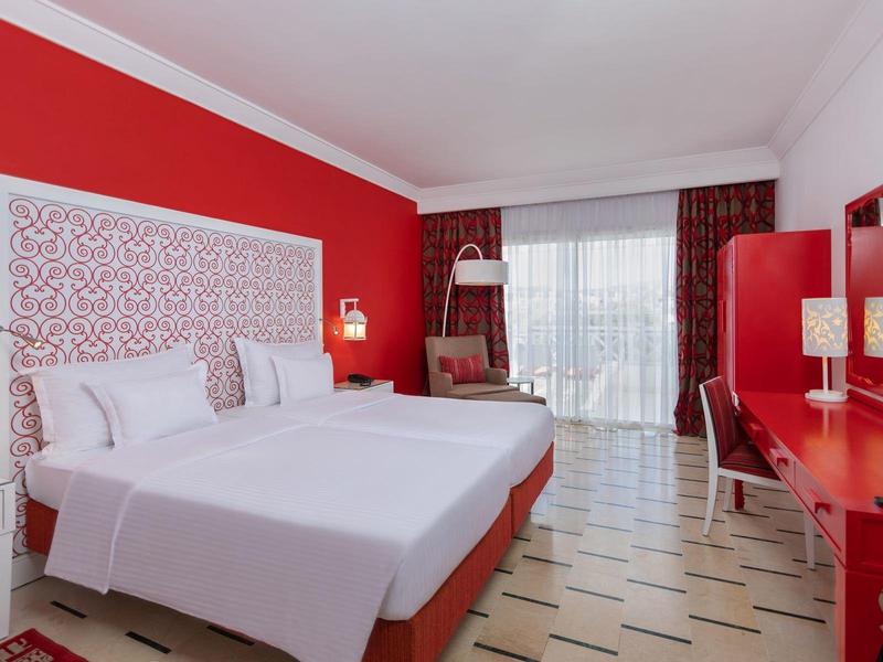 Nowoczesny pokój hotelowy z podwójnym łóżkiem, czerwonym wyposażeniem i dużym oknem z zasłonami.