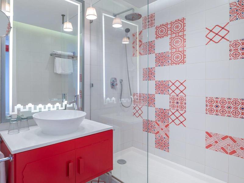 Nowoczesna łazienka z czerwoną szafką, szklaną kabiną prysznicową i czerwonymi wzorami na płytkach ściennych.