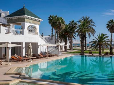 Moderner Hotelpool mit Palmen und Liegestühlen unter blauem Himmel.