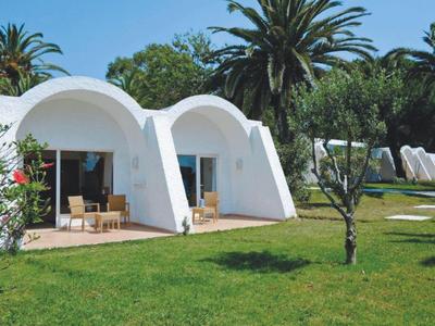 Dva bílé bungalovy s oblými střechami a terasami na zeleném trávníku s palmami v pozadí.