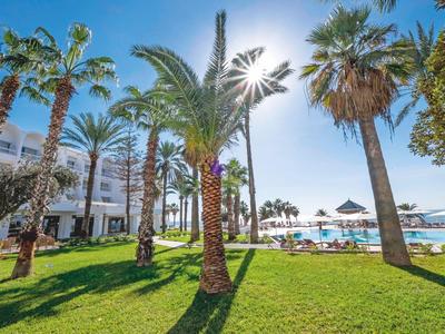 Zelený trávník s palmami před hotelem a bazénem pod jasným sluncem.