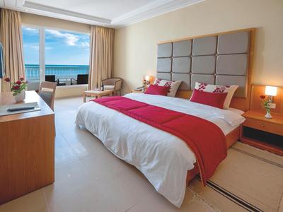 Světý hotelový pokoj s velkou postelí, červeným přehozem a výhledem na moře skrz velká okna.