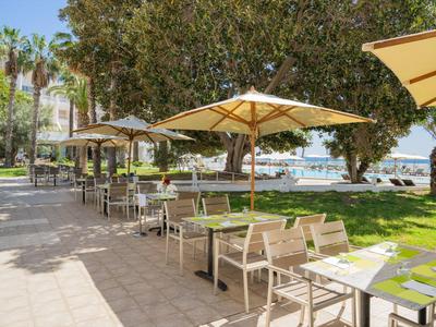 Venkovní restaurace s stoly a židlemi pod slunečníky vedle parku s výhledem na moře.