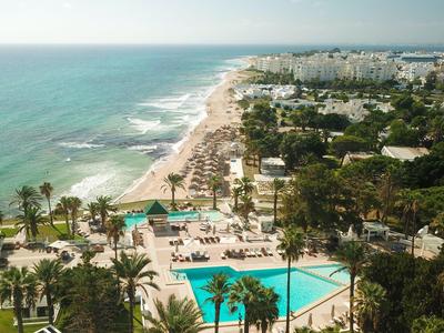 Letecký pohled na pláž s hotely, bazénem a palmami podél pobřeží.