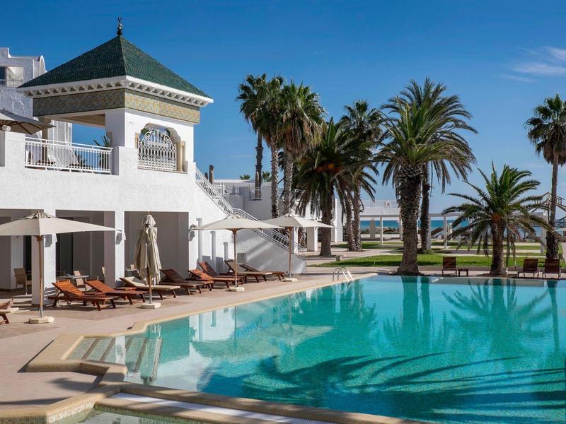 Hotel s venkovním bazénem, slunečníky a palmami pod jasně modrou oblohou.