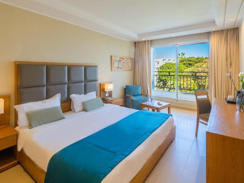 Světlý hotelový pokoj s manželskou postelí, posezením a balkonem s výhledem na zeleň.