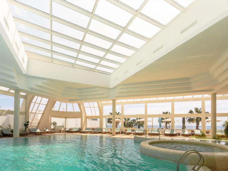 Vnitřní bazén s velkou prosklenou střechou a výhledem na palmy a moře