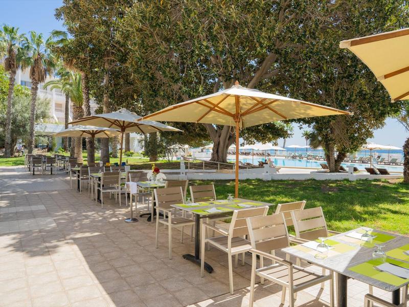 Venkovní restaurace s stoly a židlemi pod slunečníky vedle parku s výhledem na moře.