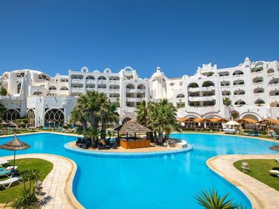 Hotelanlage mit großem Pool, Palmen, Liegestühlen und weißem Gebäude bei sonnigem Himmel.