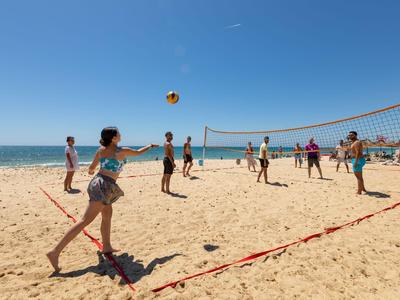 Mensen spelen beachvolleybal op zand met zee en heldere lucht op de achtergrond.
