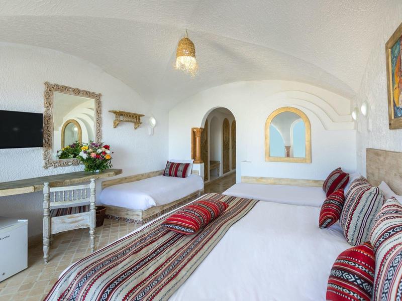 Světly hotelový pokoj s manželskou postelí, psacím stolem, židlí, zrcadlem, televizí a mini ledničkou.