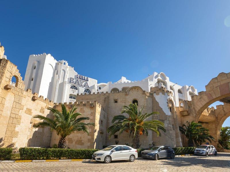 Moderní bílé budovy a oblouk na slunném pobřežním místě s palmami a zaparkovanými auty.