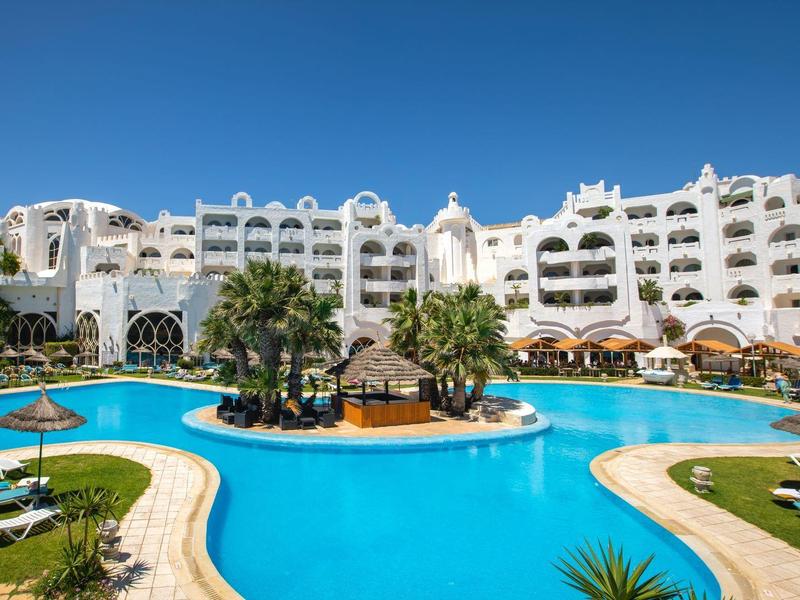 Hotelový komplex s velkým bazénem, palmami, lehátky a bílou budovou pod jasně modrou oblohou.