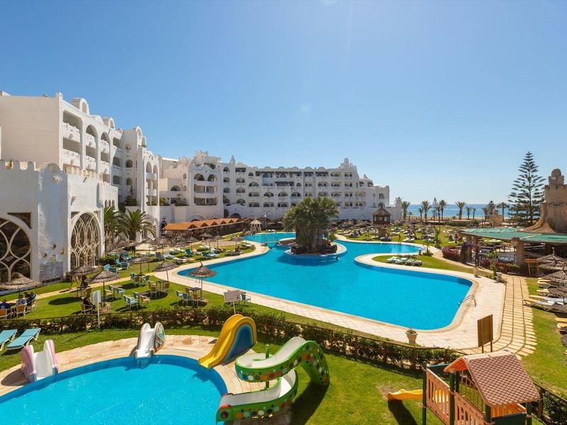 Velký hotel s několika bazény, vodními skluzavkami a palmami pod jasnou oblohou.