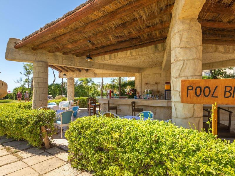 Zastřešený bar u bazénu se kamennými sloupy, obklopen keři a venkovním posezením.