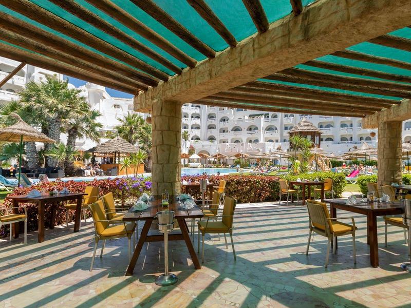 Hotelová venkovní terasa s stolky a židlemi pod sluneční clonou.