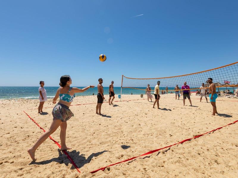 Lidé hrají volejbal na písečné pláži za jasné oblohy a klidného moře.