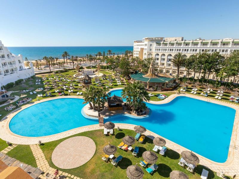 Velký bazén s deštníky mezi dvěma hotelovými budovami a výhledem na moře.