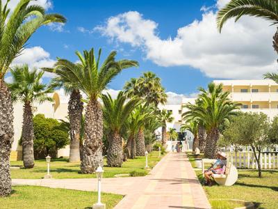 Široká zahradní cesta lemovaná palmami a zelenými trávníky vedoucí k budově resortu za slunečného počasí.