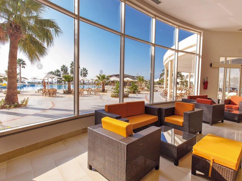 Lounge dell'hotel luminosa con grandi finestre che affacciano su palme e mare.