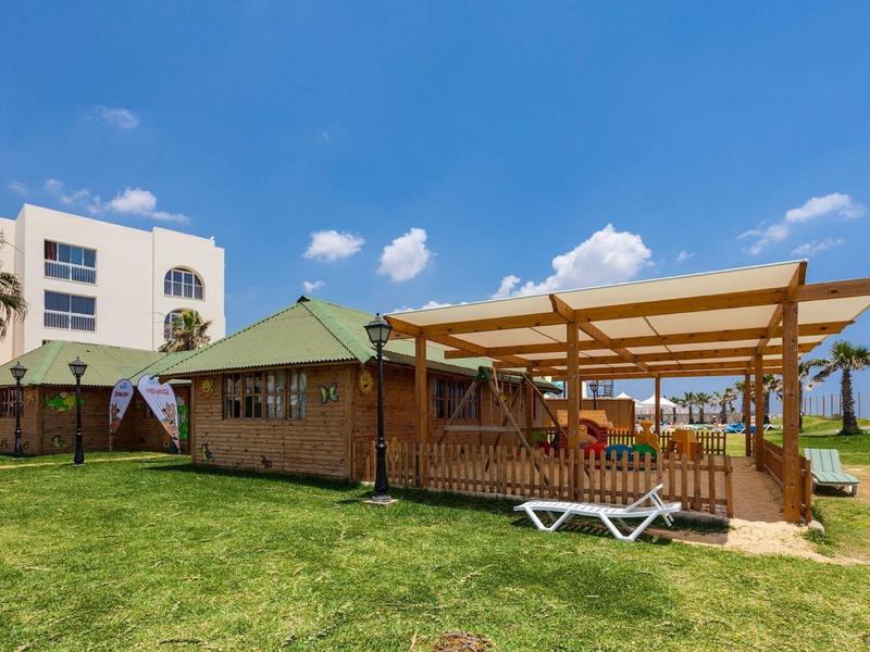 Grande edificio in legno con terrazza coperta e lettini davanti a un hotel bianco sotto un cielo azzurro.