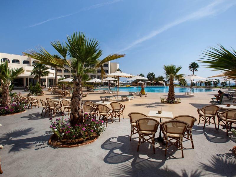Terrazza dell'hotel con tavoli e sedie accanto a una piscina e palme in una giornata di sole.