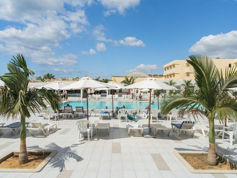 Area esterna luminosa dell'hotel con piscina, palme e ombrelloni bianchi sotto un cielo azzurro.