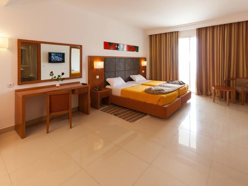 Camera d'hotel luminosa con letto matrimoniale, scrivania, sedie e grandi finestre con tende.